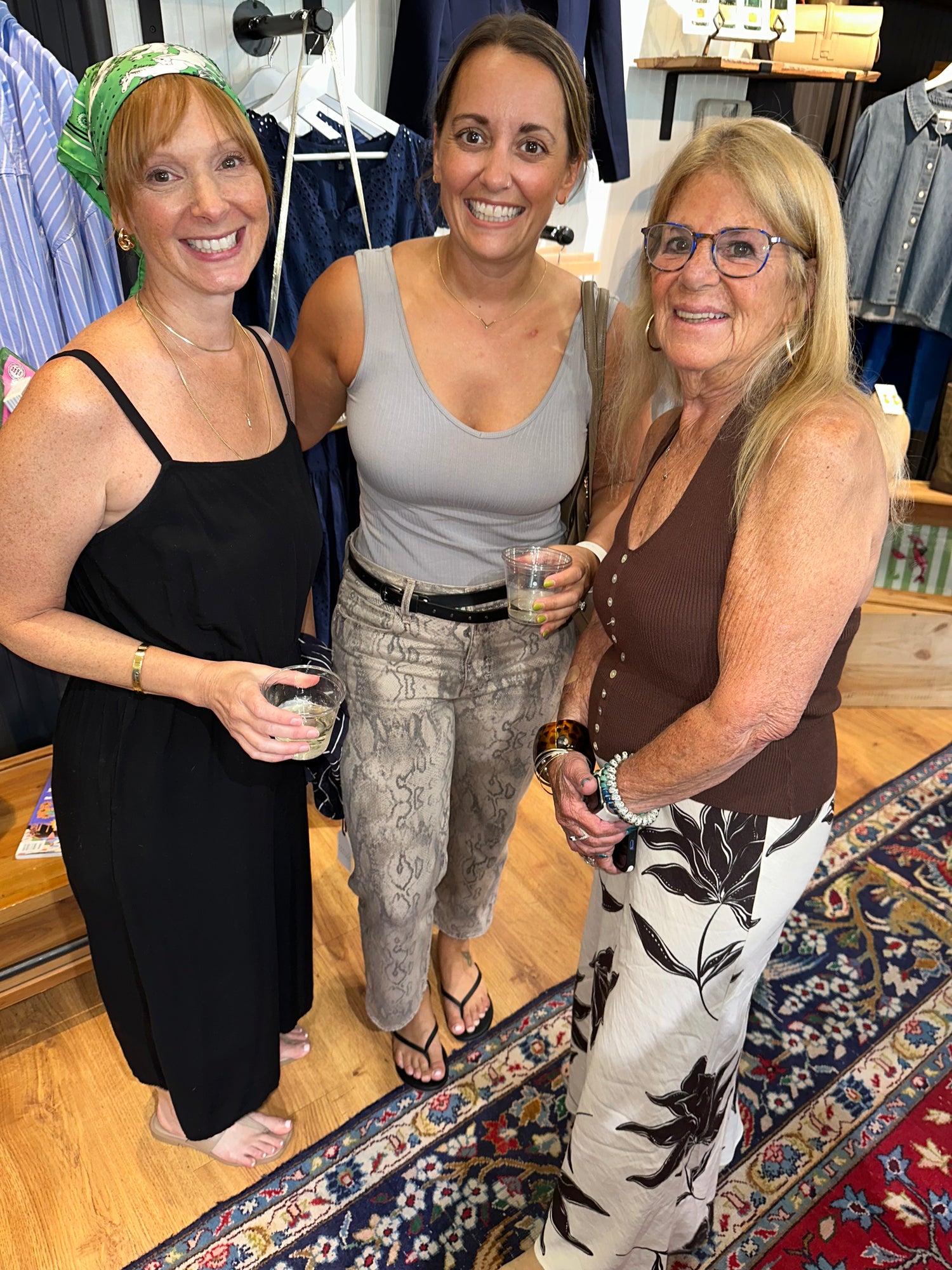 Three women smiling and posing together inside a clothing boutique. The woman on the left wears a black sleeveless dress, the middle woman wears a light gray tank top and patterned pants, and the woman on the right wears a brown tank top and white pants with a black floral print.
