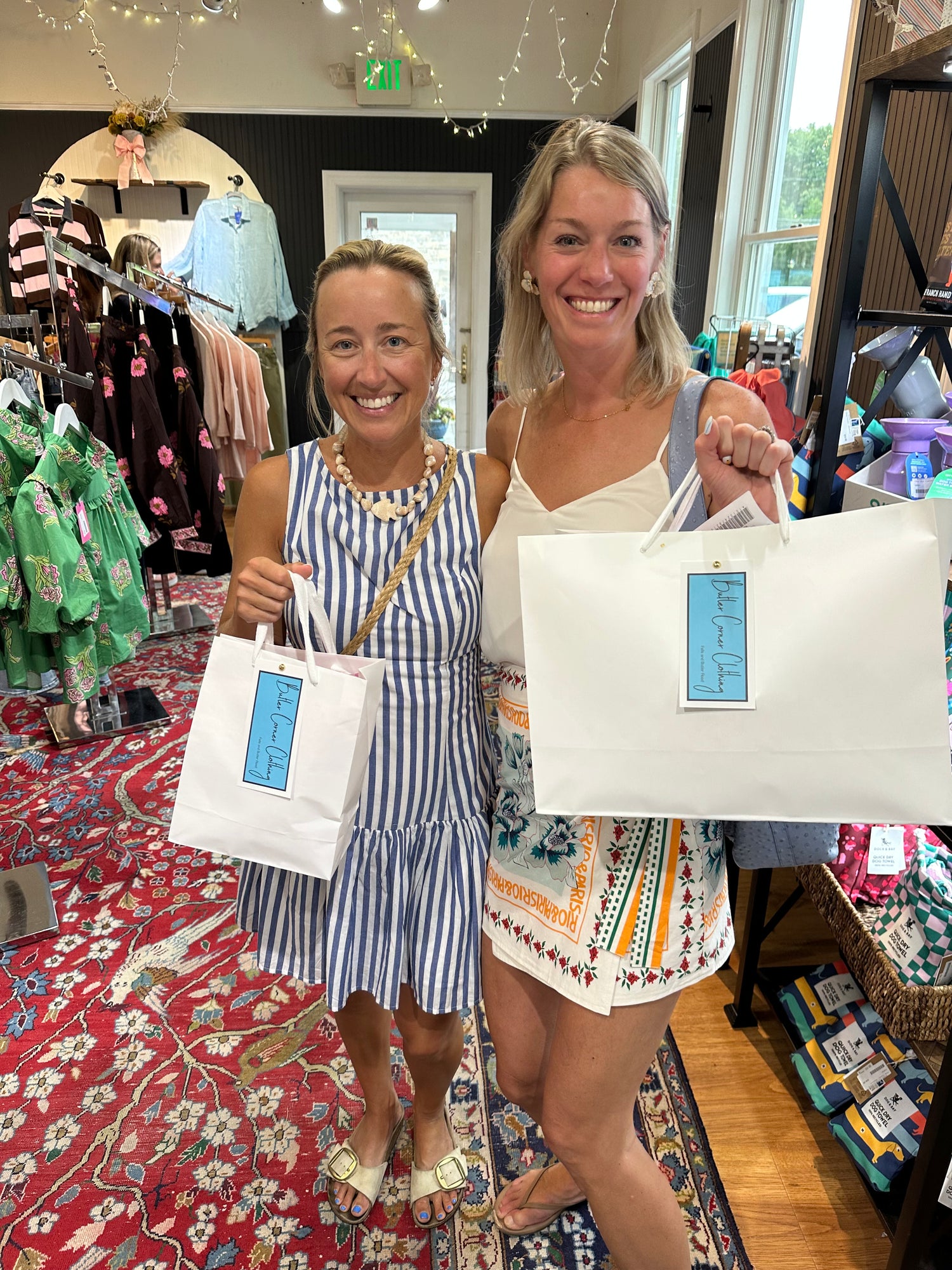 Two women smiling inside a boutique shop, each holding a white paper shopping bag with a blue 'Butler Corner Clothing' tag. The woman on the left wears a blue and white striped dress, and the woman on the right wears a white top with a multicolor patterned skirt.